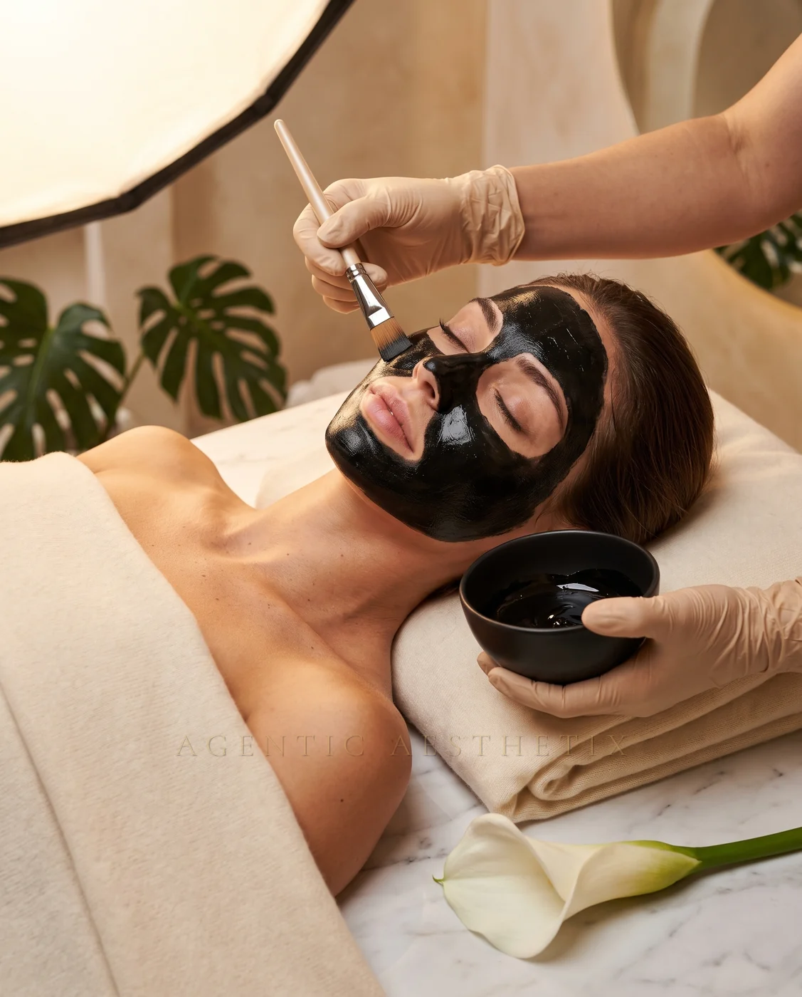 Luxury black charcoal facial mask treatment, practitioner applying mask with brush on treatment bed with marble and tropical plants